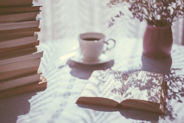 a sun-dappled table covered in a pile of books with a cup of tea