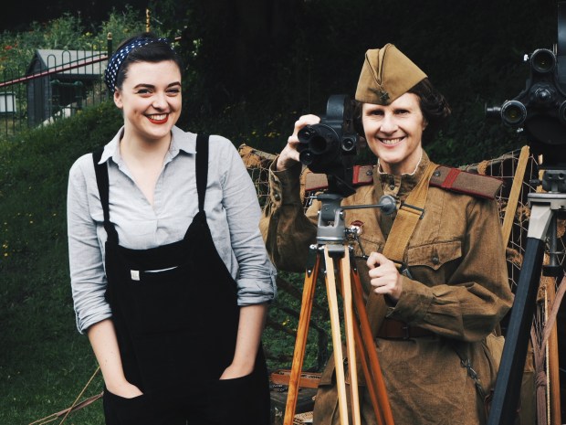 A girl in vintage dungarees next to a woman in WW2 soviet uniform with a video camera