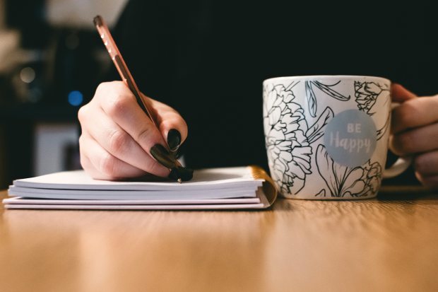Woman's hands writing in a notebook with black nail polish and a cup with 'be happy' on it