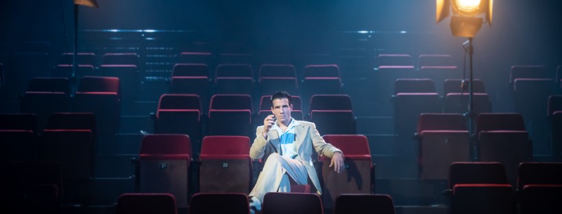 a man in a light-coloured suit lounges in a theatre seat