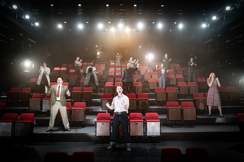 a theatre cast stand, spaced out, in among theatre seats