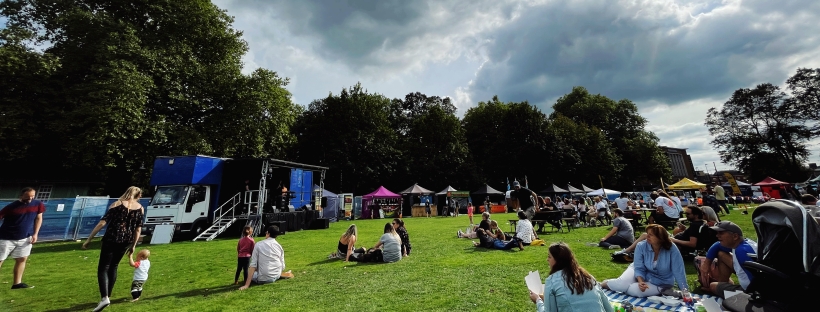 People sitting on a field surrounded by festival stalls, watching a stage