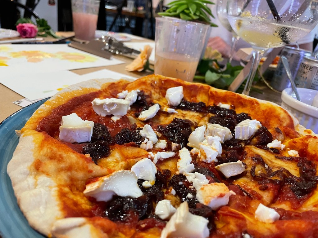 A goat's cheese and onion marmalade pizza; behind it, you can see a glass of paint-filled water, a gin and tonic, and a blurred painting